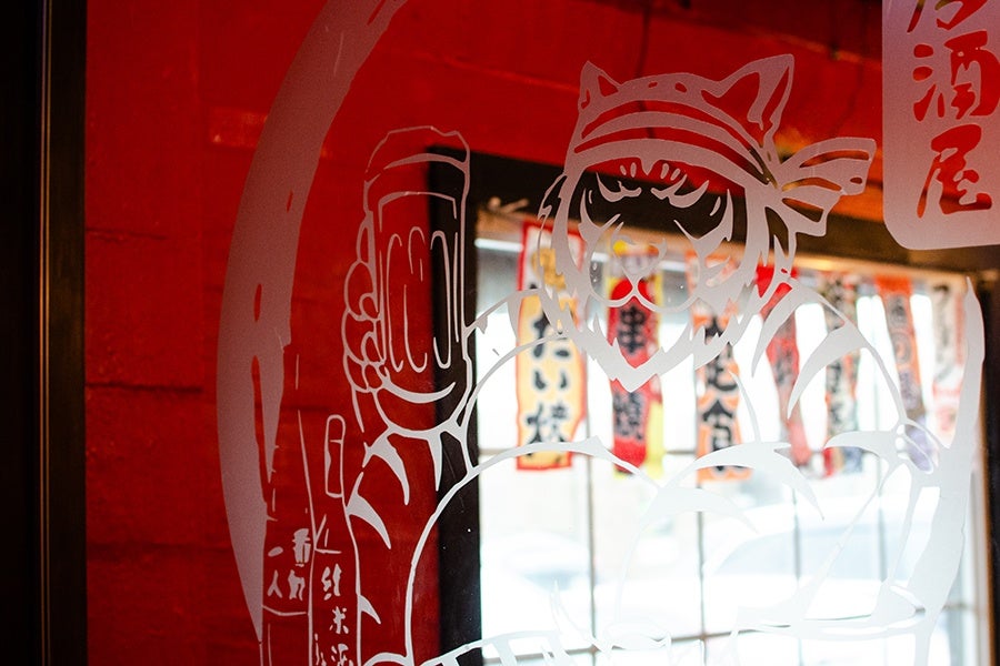 A cartoony fat tiger holding a mug of beer decorates a glass partition inside a restaurant, with a red wall and window visible behind and small colorful flags hanging above the window.