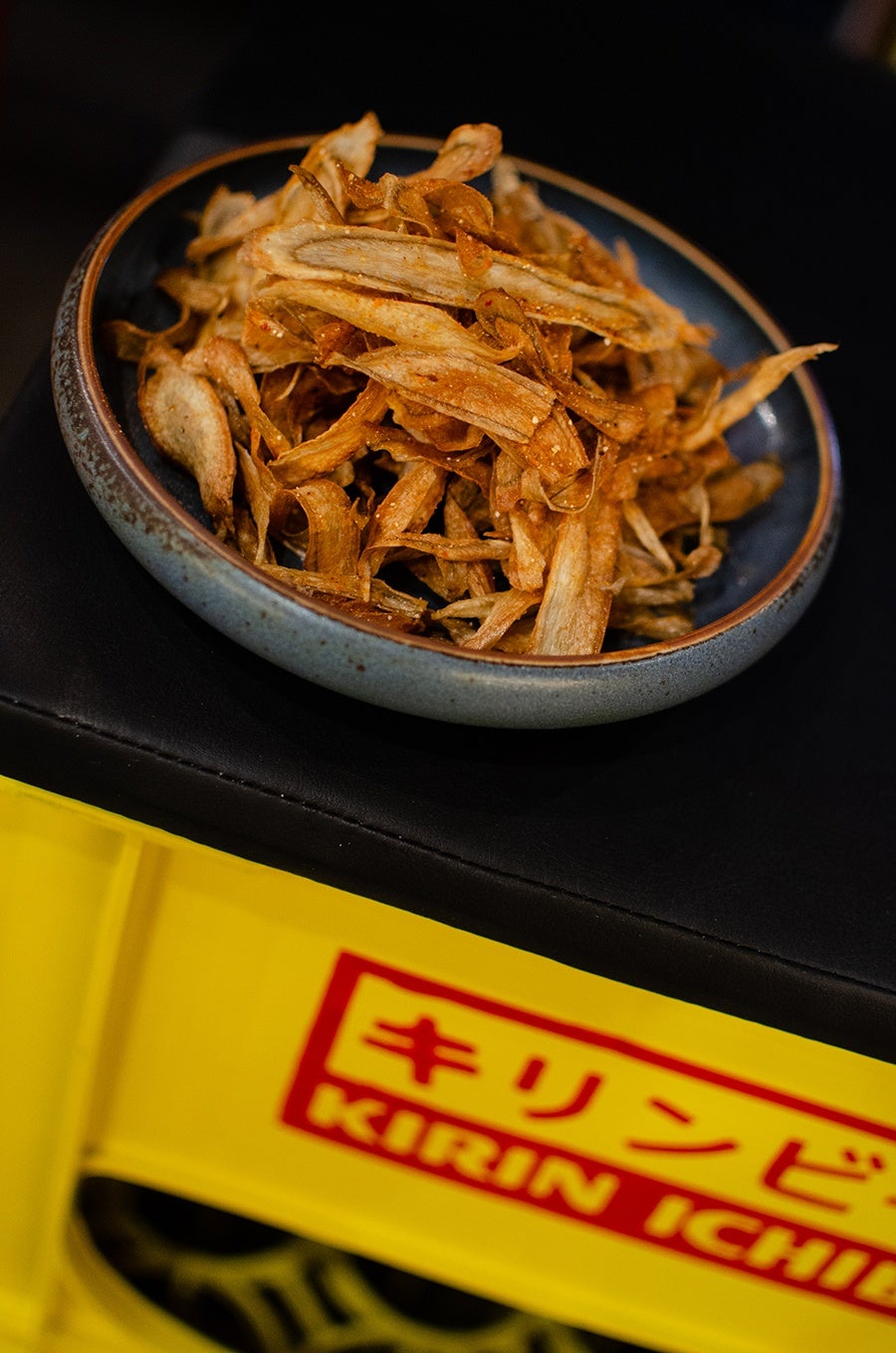 Crispy fried slivers of burdock root are displayed in a blue bowl atop a yellow Japanese beer crate.