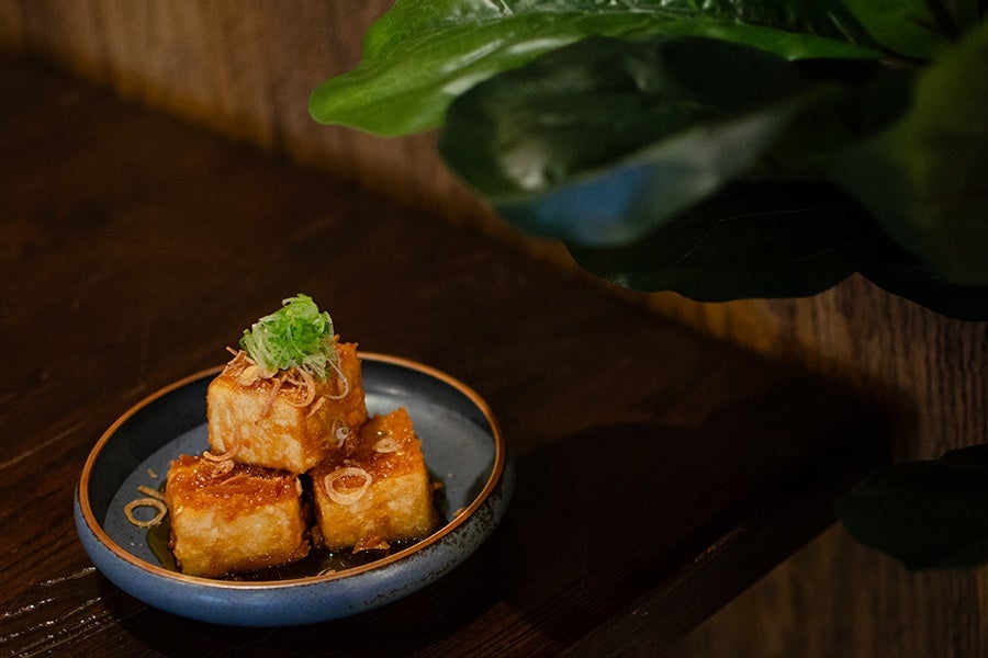 Crispy fried tofu in a sticky brown sauce sit on a blue plate on a table near some faux greenery.