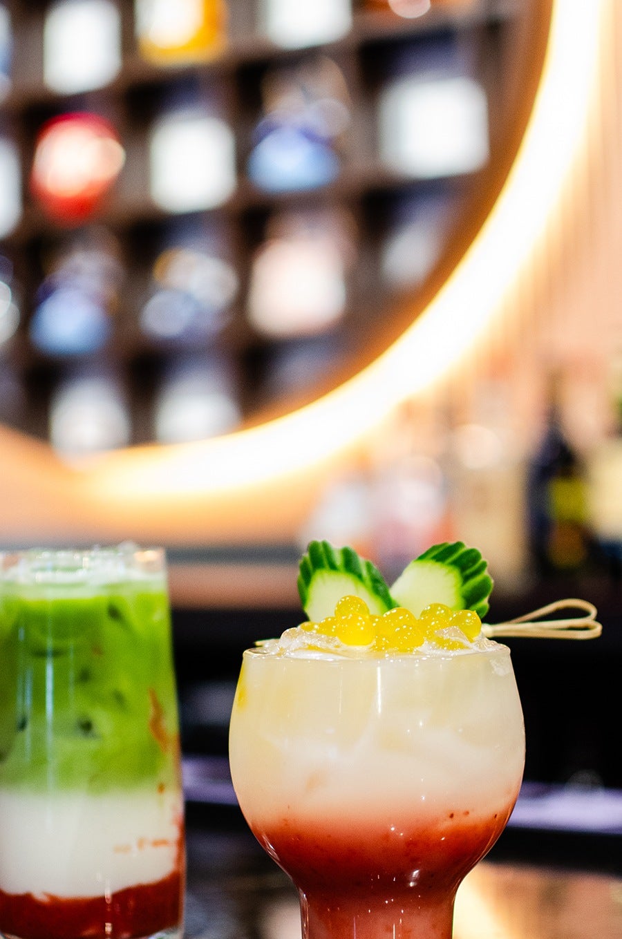 Two colorful drinks, one topped with yellow boba and cucumbers, sit on a bar.