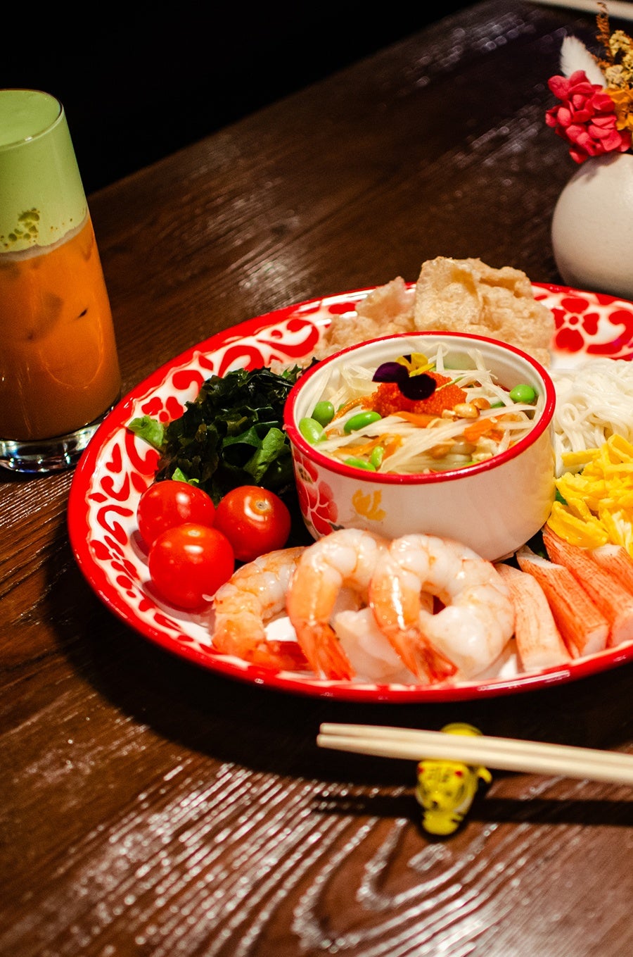 A big red and white plate is filled neatly with a Thai-style papaya salad and various accents, some Thai and some Japanese, including noodles, tomatoes, crispy pork rinds, and more.