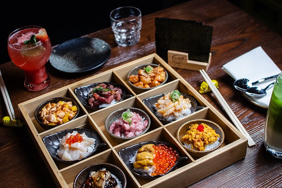 A 3x3 wooden box is filled with different Thai-Japanese fusion hand roll fillings. To the side, chopsticks rest on small tiger figures.