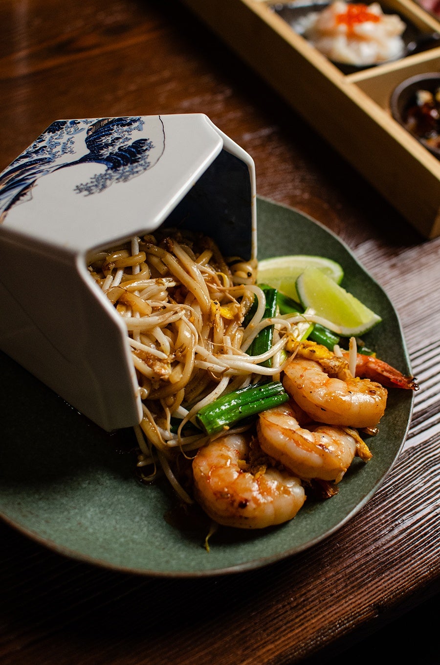 Pad thai with shrimp spills out of a ceramic container that looks like a takeout box decorated with Hokusai's wave.