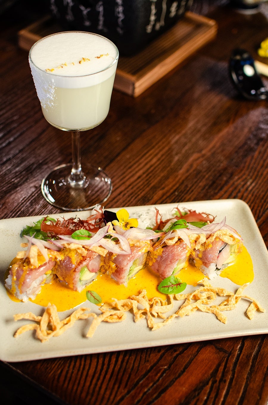 A pale yellow cocktail with a foam top sits next to rare beef-topped maki sitting in a bright yellow sauce, with flower and crispy noodle garnish.