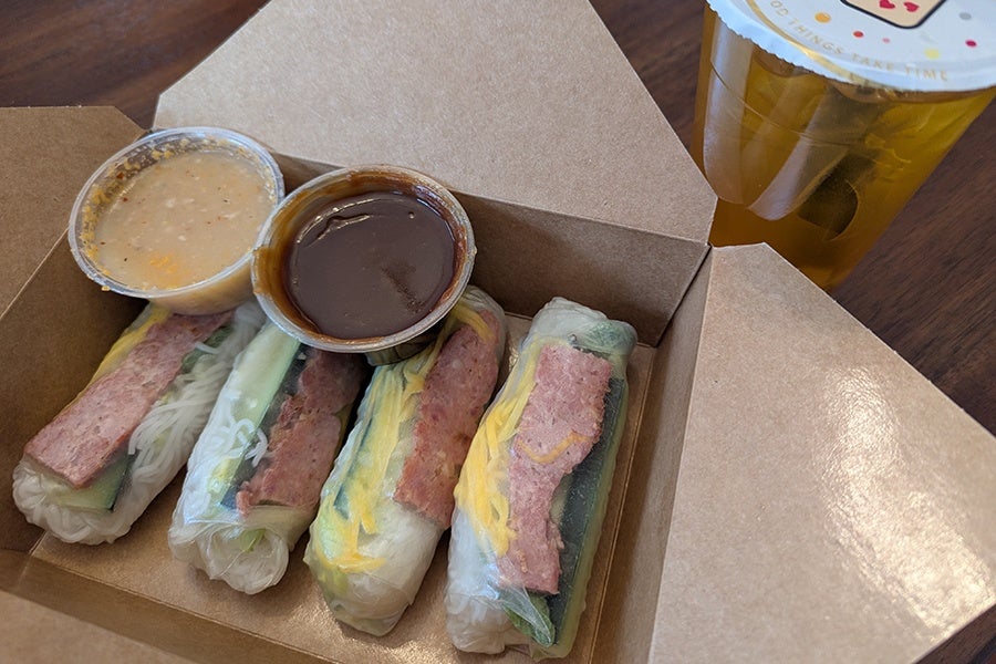Four rice paper rolls are lined up in a takeout box, each stuffed with pink sausage and vermicelli.
