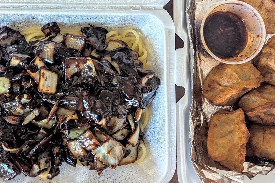Styrofoam takeout containers hold long wheat noodles in a shiny black bean sauce and fried dumplings.