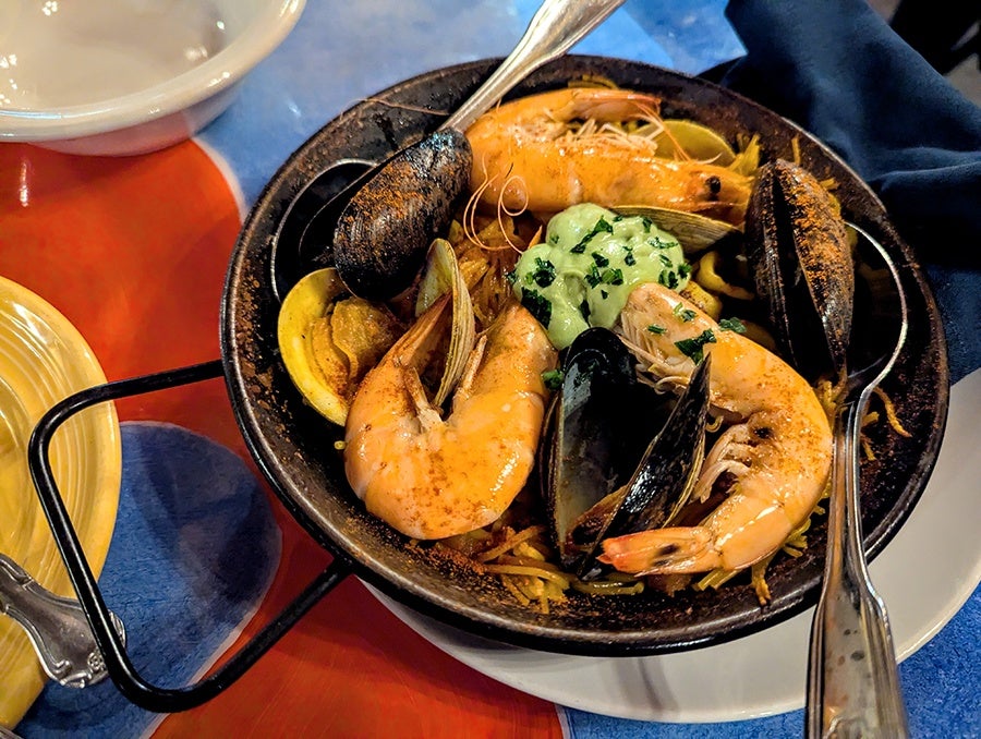 Three large shrimp and some mussels perch atop thin noodles in a small paella-style pan.