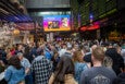 A crowd of people pack into a food hall with "Big Queer Food Fest" branding on a giant projection screen overhead.