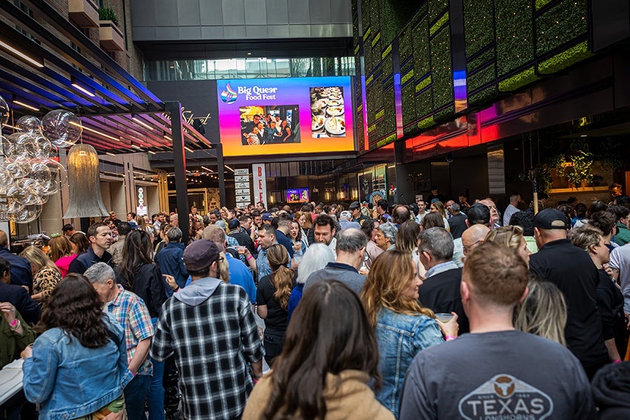 A crowd of people pack into a food hall with "Big Queer Food Fest" branding on a giant projection screen overhead.