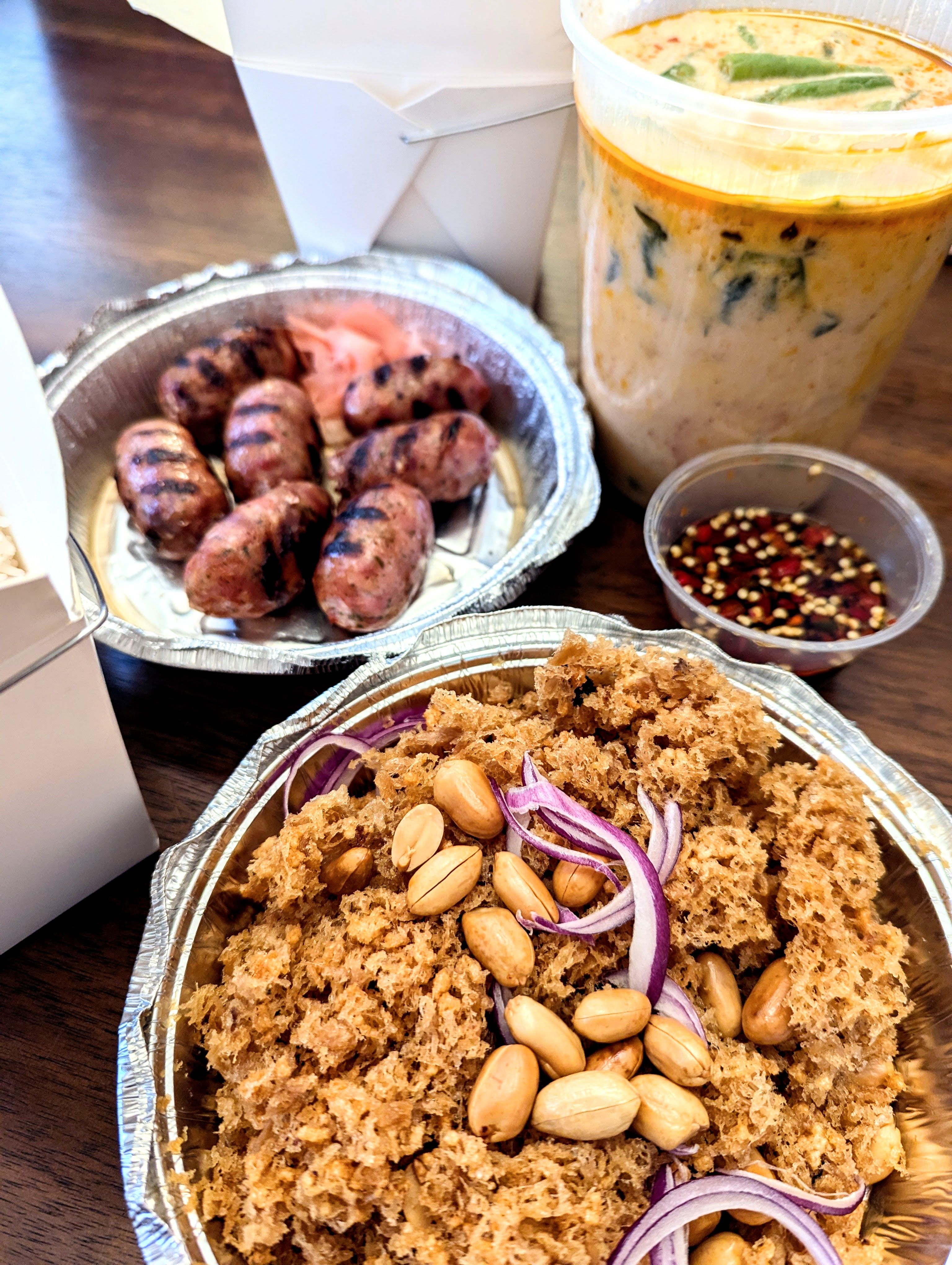 Thai food in delivery containers, including small plump sausages, a cloud of fried fish topped with peanuts, and a big container of pale orange curry.