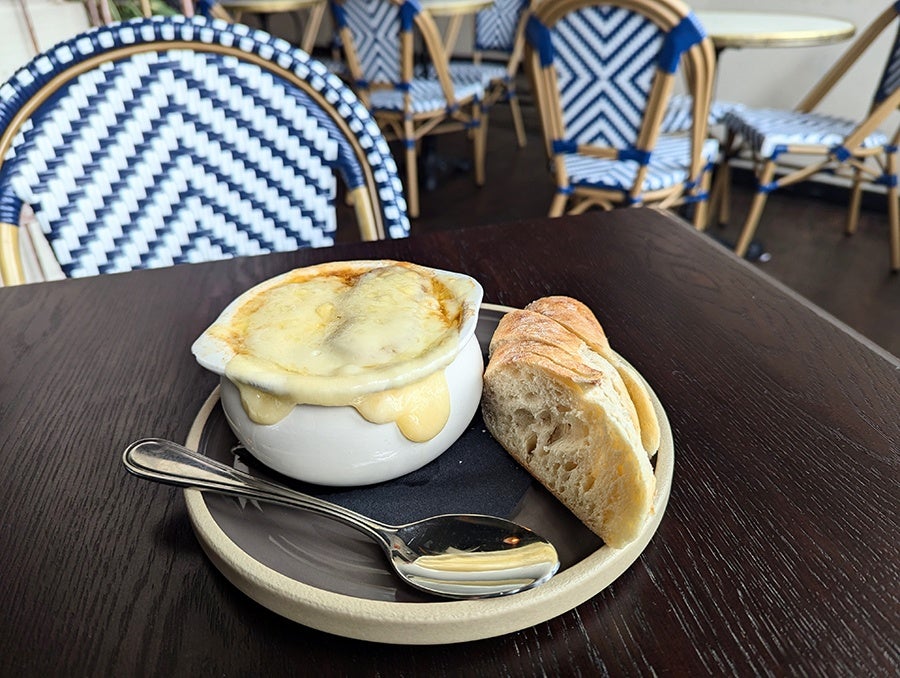 A bowl of French onion soup, cheese dripping down the sides, is accompanied by a sliced baguette and sits on a cafe table.