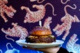 A hand holds up a burger on a sesame bun with pickles, cheese, and sauce in front of tiger wallpaper.