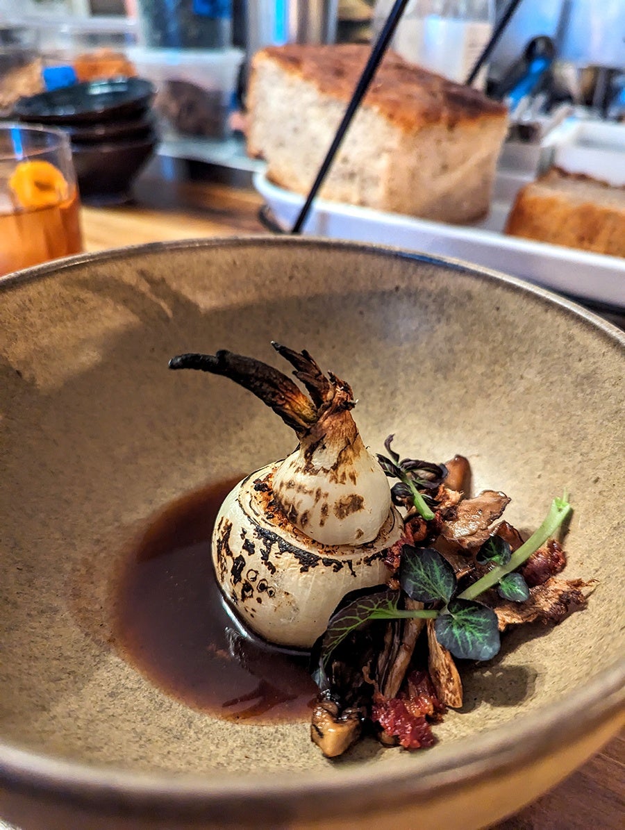 A fine-dining dish with an onion and some vegetables plated prettily at a chef's counter.