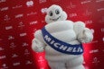 A person in an inflatable Michelin Man suit stands in front of a red backdrop covered with brand names at an event, giving two thumbs up.