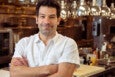 A chef in a white collared shirt stands, arms crossed, smiling at the camera in front of a restaurant kitchen.