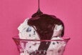 Hot fudge is poured over a scoop of white ice cream with chocolate chips in a glass bowl, photographed on a pink background.