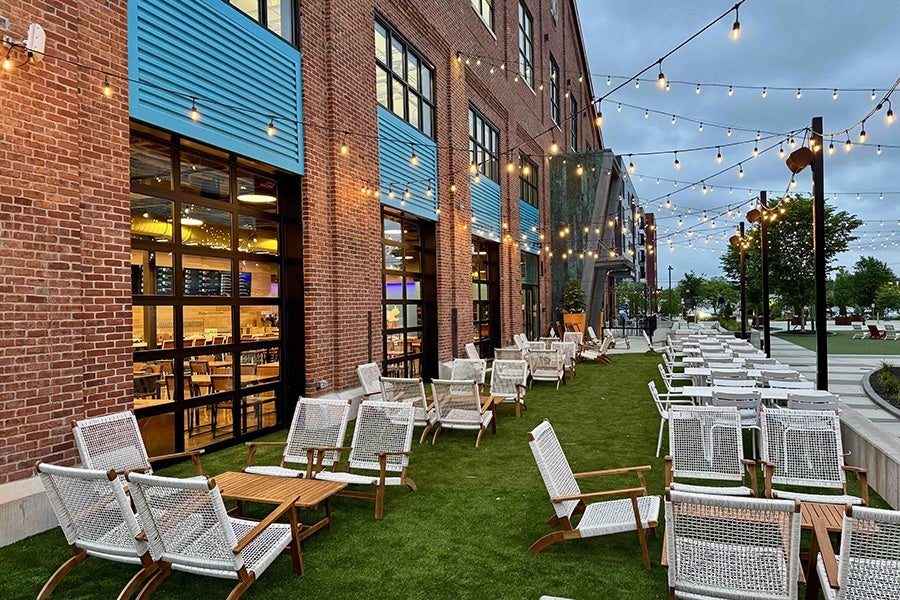 Lounge chairs and low tables are set up on a strip of turf next to a brick building with big windows.