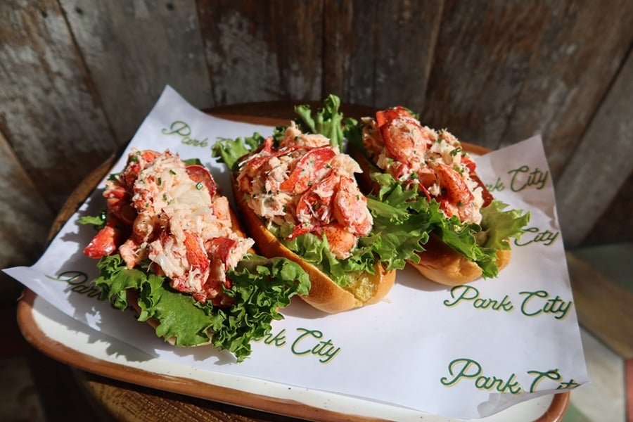 Three lobster rolls with mayo and lettuce are served in hot dog buns, sitting on a piece of paper that says Park City all over it.