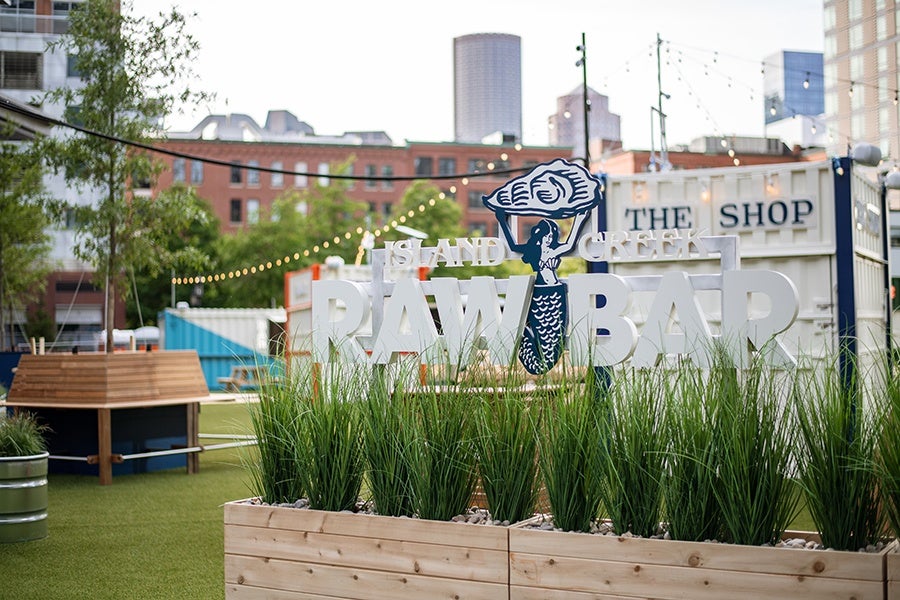 Signage reading Island Creek Raw Bar features a mermaid holding up an oyster. The sign is situated in front of a turf-lined outdoor dining area.