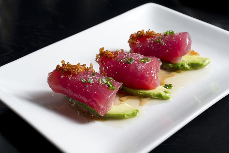 Three slices of raw tuna are rolled up and served on sliced avocado, garnished with herbs.