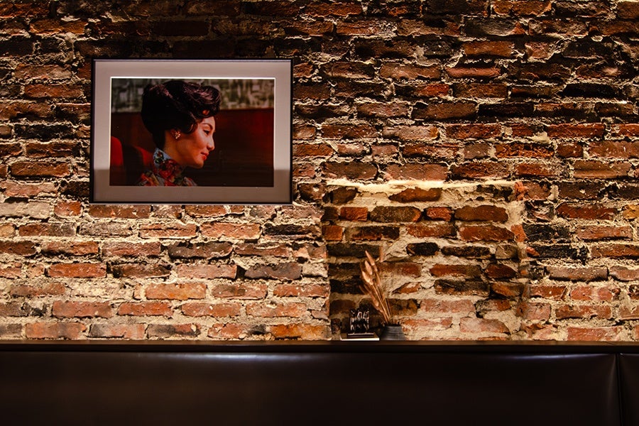 A framed photograph of a woman is displayed on an exposed brick wall over a dark brown banquette.