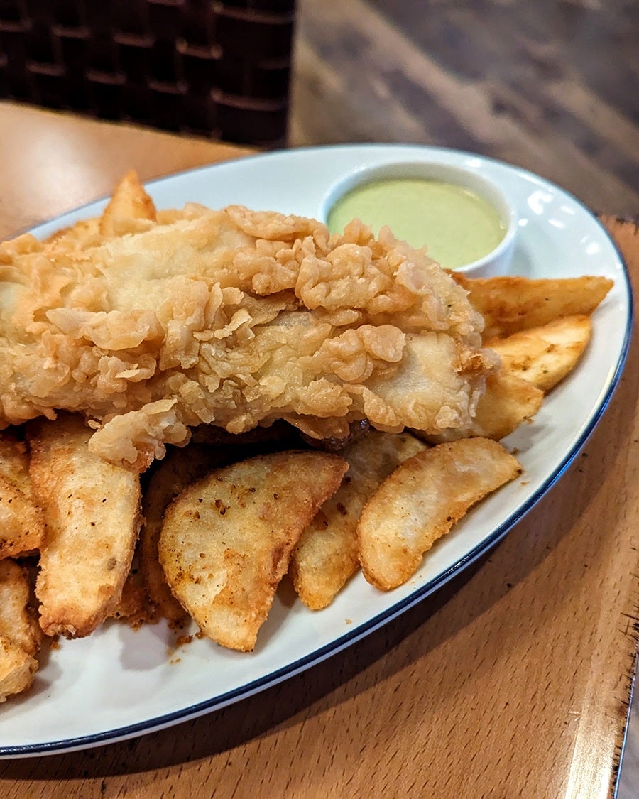 A big piece of crispy fried fish sits atop spice-dusted big potato wedges with a creamy pale green sauce on the side.