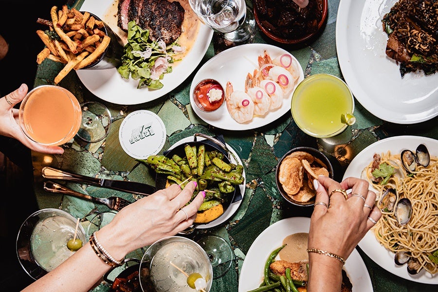 Hands pick up food and drink from a green table covered with linguine and clams, steak frites, shrimp cocktail, and more.