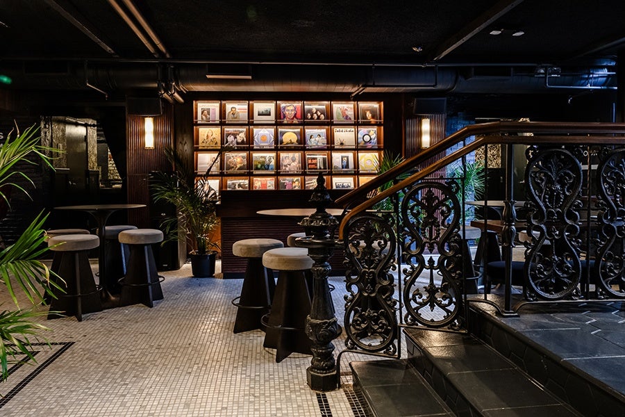 Stairs with an ornate black railing descend into a white-tiled, dimly lit restaurant space, featuring a wall of vinyl records.