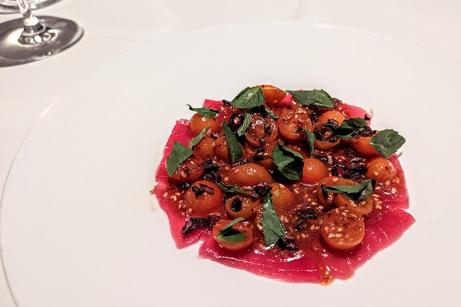 Thinly sliced raw tuna is arranged on a plate under sliced tomatoes and herbs.