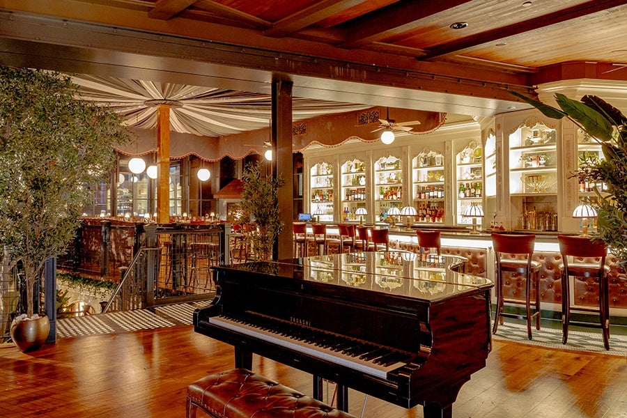Interior of a restaurant features a grand piano, a bar upholstered in brown leather with a white wooden backbar, and a pale green-and-white-striped canopy over the dining room.