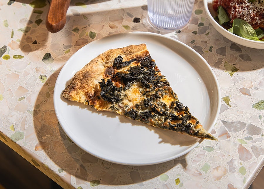 A slice of pizza topped with braised greens sits on a white plate on a terrazzo table.