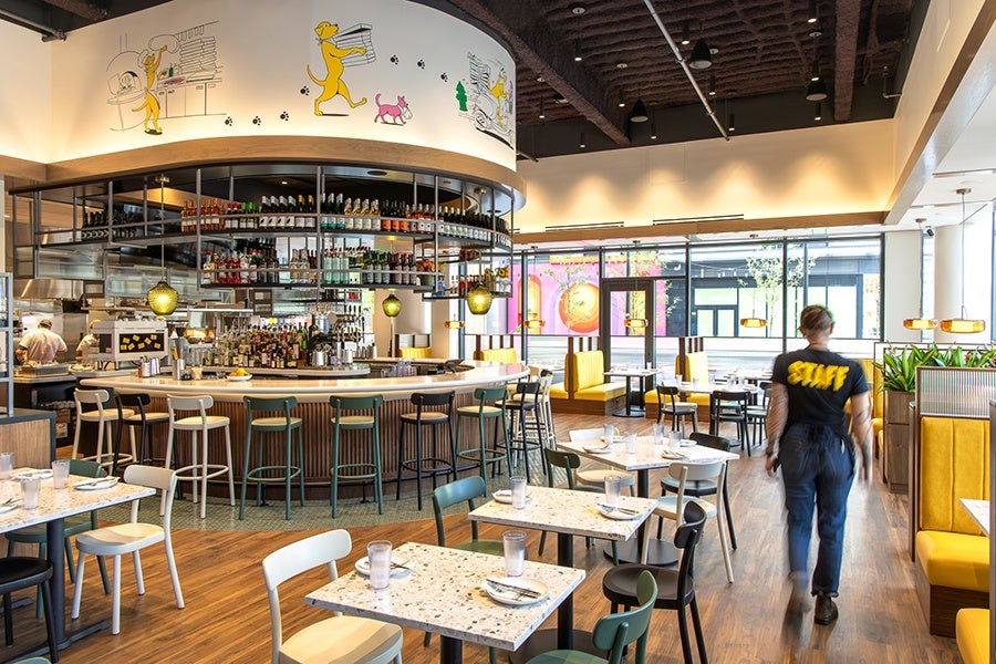 Somewhat casual, full-service restaurant interior with a big u-shaped bar, a cartoony mural of a dog making and delivery pizzas, yellow banquettes, and terrazzo tables.