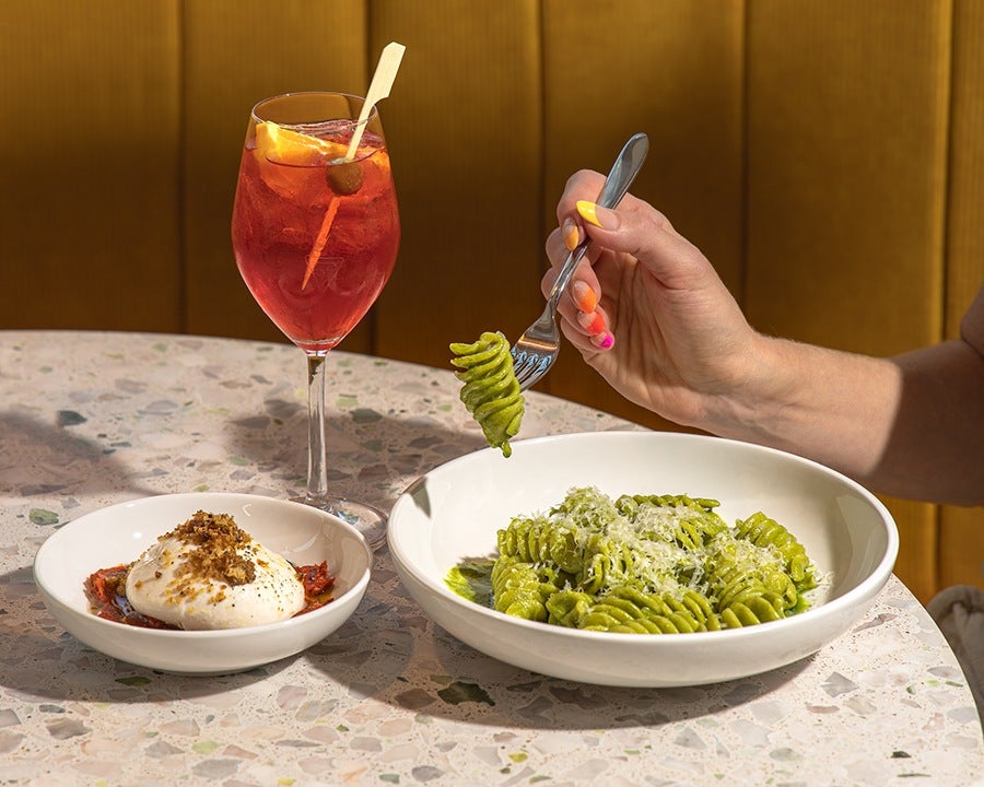 A hand holds a fork with a piece of twirly pasta on it, in a green sauce. A spritz with an orange slice and green olive accompanies the meal.