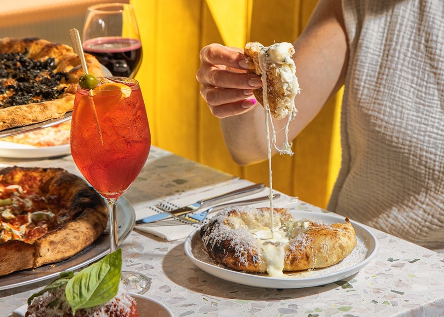A hand pulls a piece of cheesy bread up from a bigger portion, with a spritz, red wine, and pizzas visible in the background.