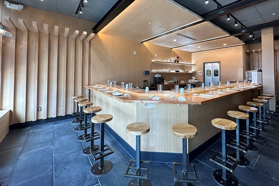 An empty restaurant interior shows a light wooden sushi bar and dark gray tiled floor.