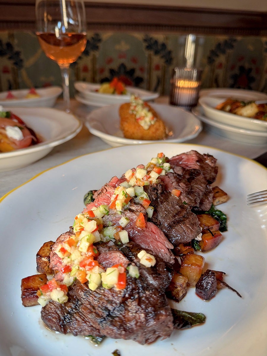 Sliced steak is topped with chopped veggies, with other dishes and a glass of wine visible in the background.