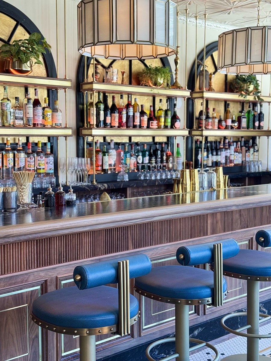 Blue leather barstools sit in front of a dark wooden bar with a black marble top and gold-accented backbar shelving.