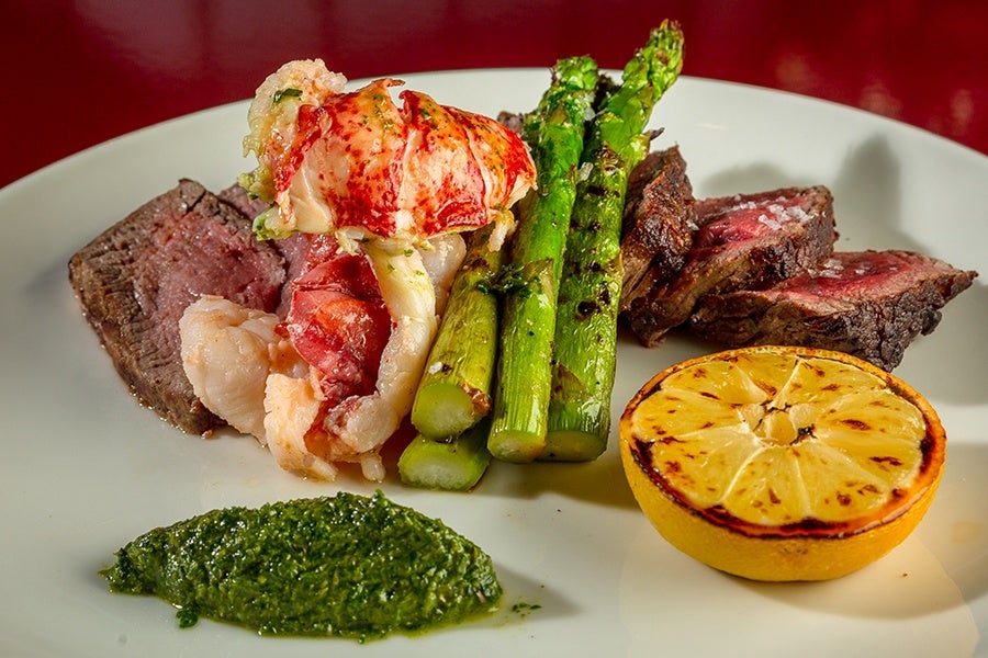 Sliced steak is served with asparagus, lobster, charred lemon, and an herby green sauce.