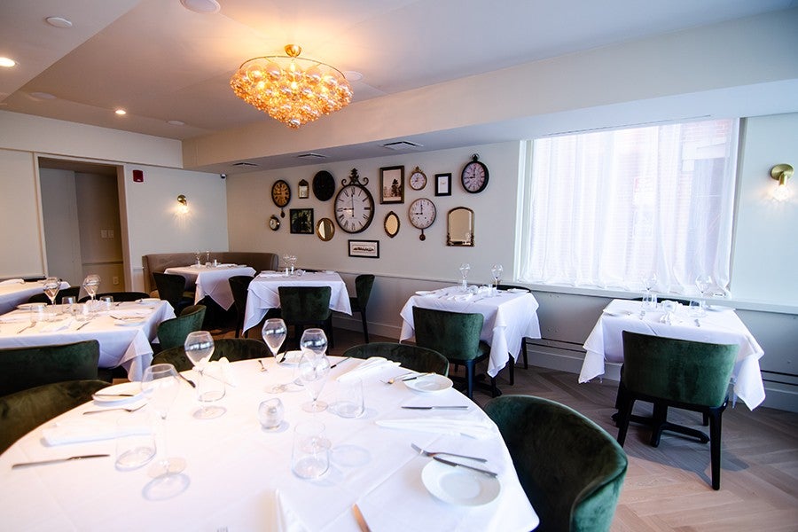 A restaurant dining room featuring dark green velvet seating, a wall of antique-looking clocks all turned to 9 o'clock, and white tablecloths.