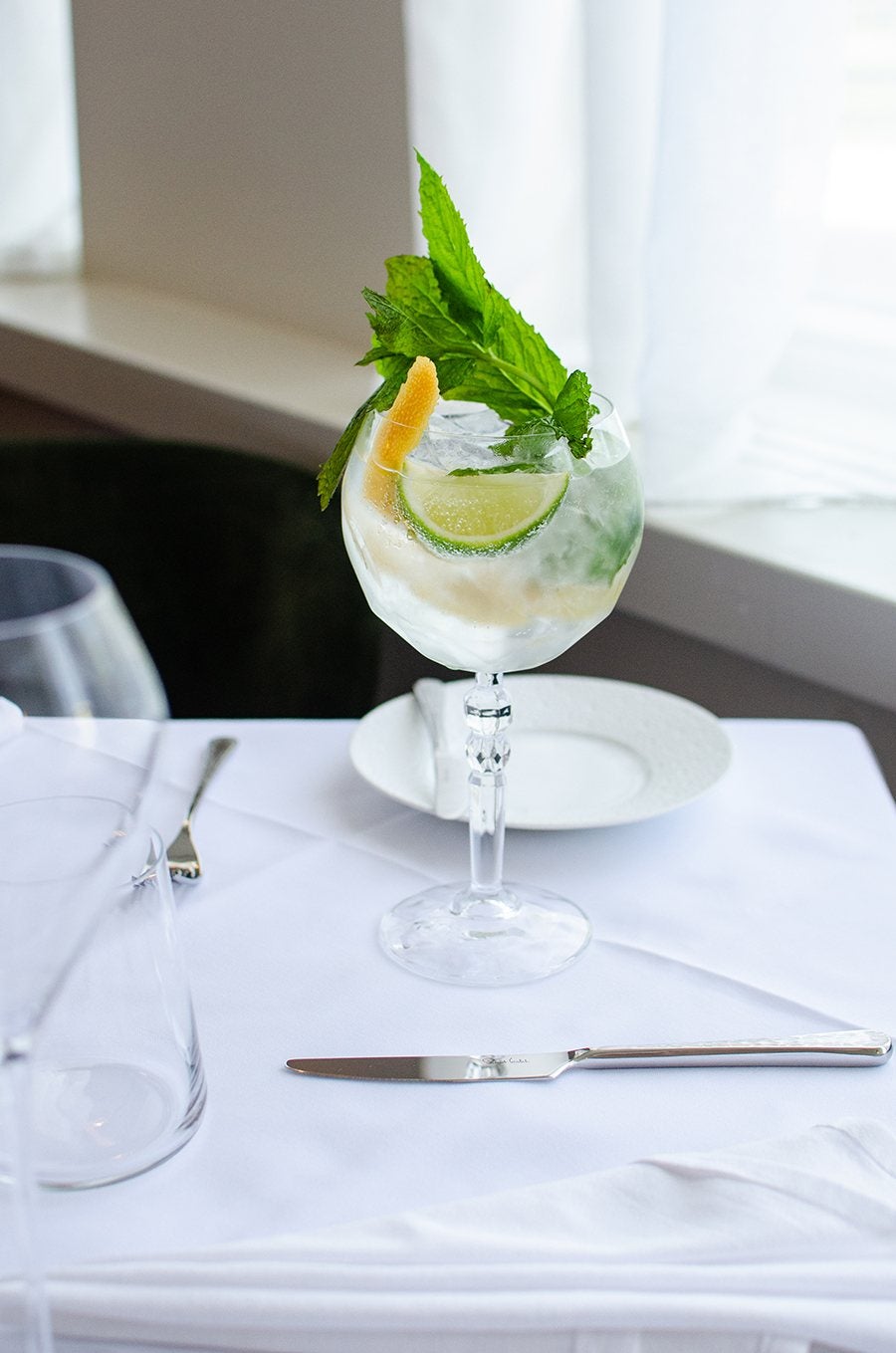 A gin and tonic in a big goblet-style glass, garnished with mint and citrus, photographed on a white tablecloth-covered table.
