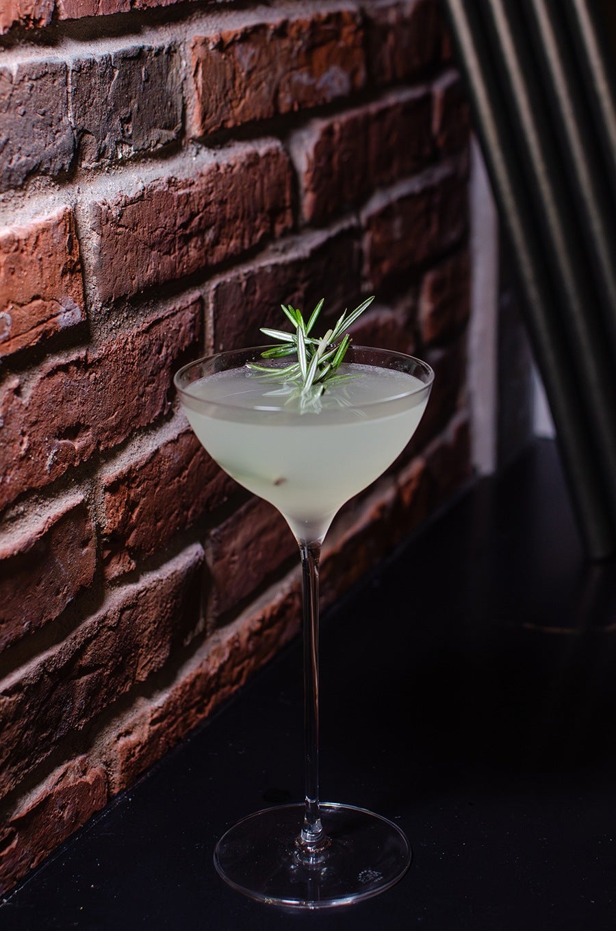 A pale yellow cocktail garnished with rosemary, photographed next to a brick wall.