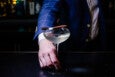 A bartender in a blue jacket holds the stem of a glass holding a pale white-yellow cocktail topped with a spoon full of caviar.