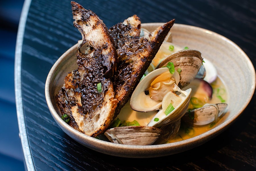 Steamed clams sit in broth in a shallow bowl, garnished with toast slathered with a shiny black spread.