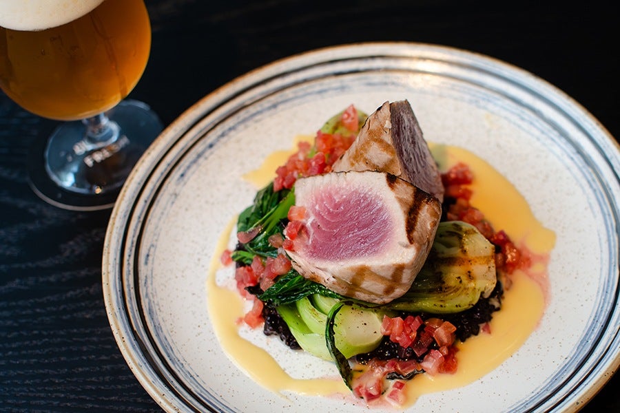Seared chunks of tuna sit atop bok choy, chopped tomatoes, black rice, and a buttery sauce, accompanied by a glass of beer.