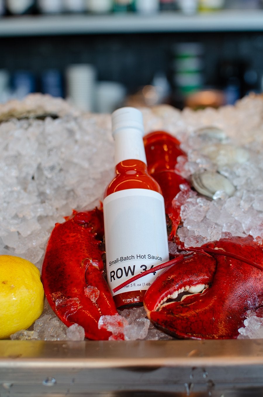 A bottle of hot sauce sits in the claws of a lobster on a raw bar, next to a lemon.