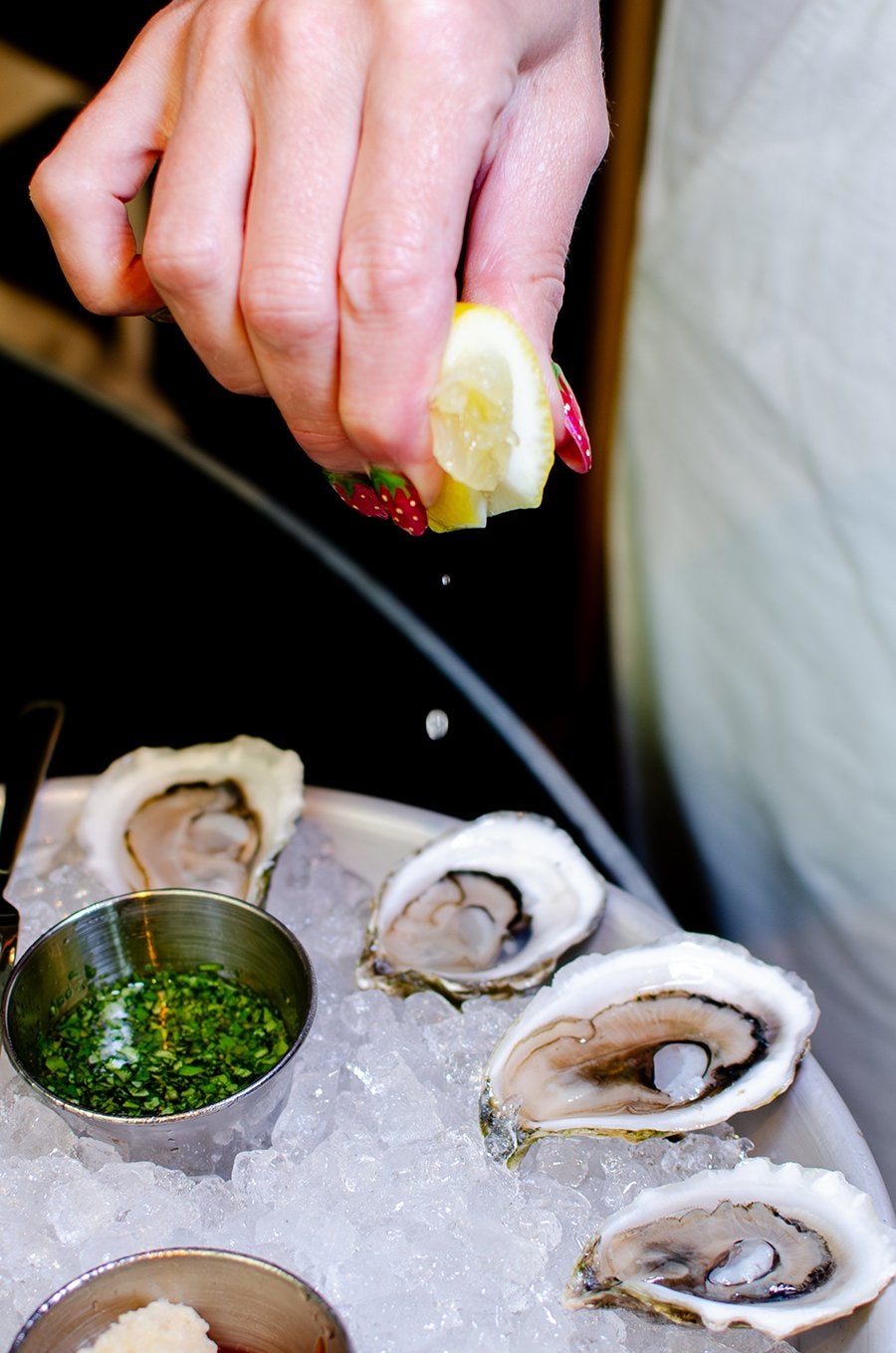 A hand squeezes a lemon wedge above a tray of oysters on the half shell on ice.