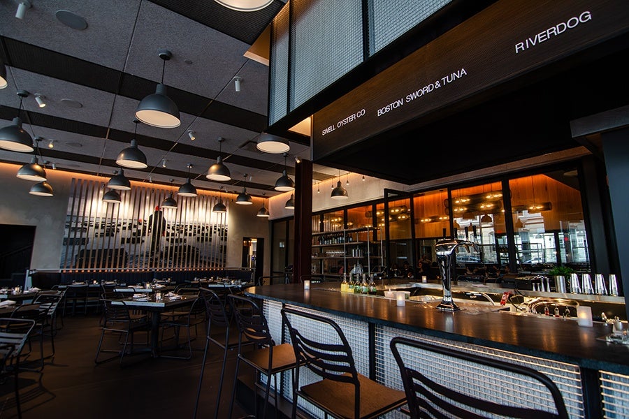 A restaurant interior features a black menu board over bar seating with the names of seafood companies, and a large photo of an oyster farm is visible in the back of the dining room.