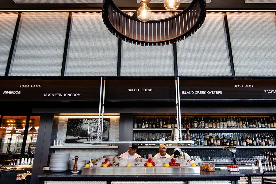 Three employees of a seafood restaurant stand working behind a raw bar under a black menu board that reads "super fresh" and lists the names of seafood companies.