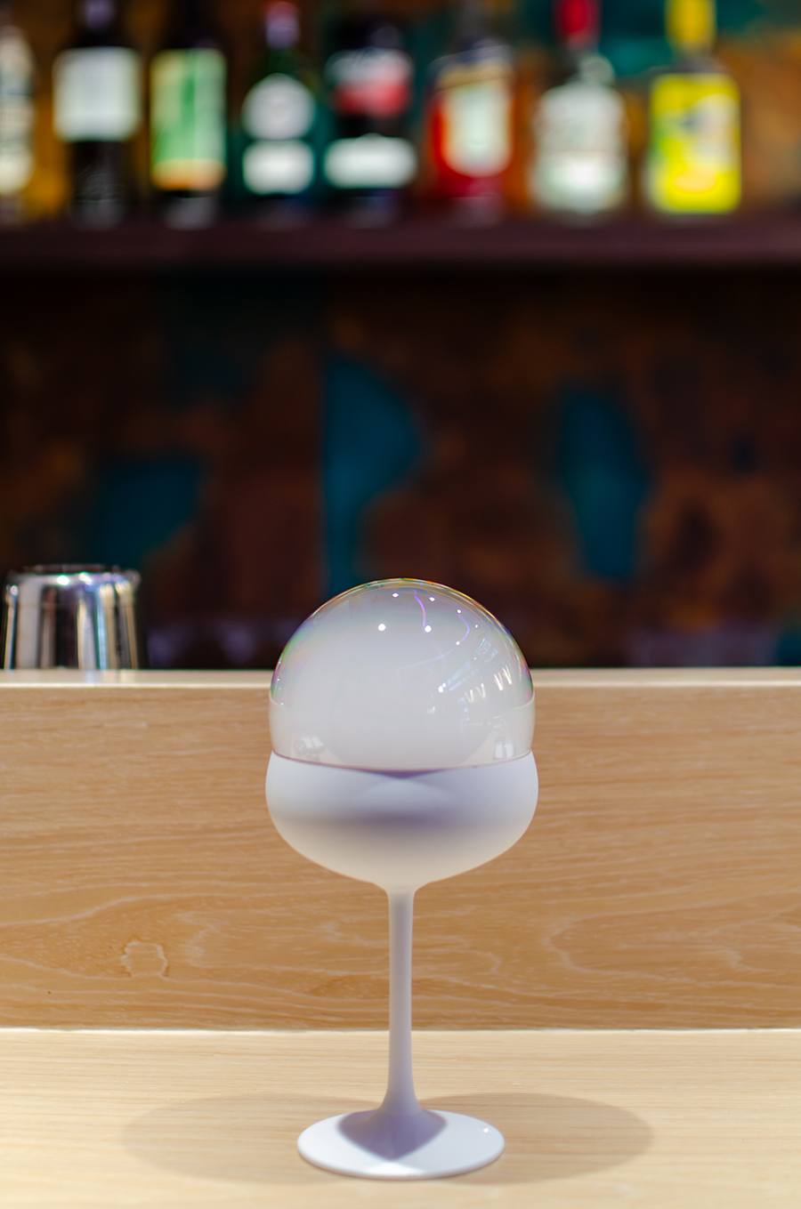 A cocktail topped with a smoky bubble sits on a light wooden bar.