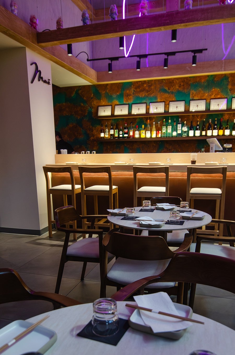 Interior of a restaurant, with the name Mai in cursive on the wall above a small light wooden bar. Labubus line a rafter above the bar.